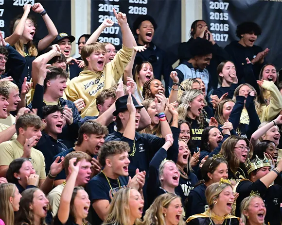 A large group of excited high school students cheering enthusiastically in a gymnasium with championship banners in the background.