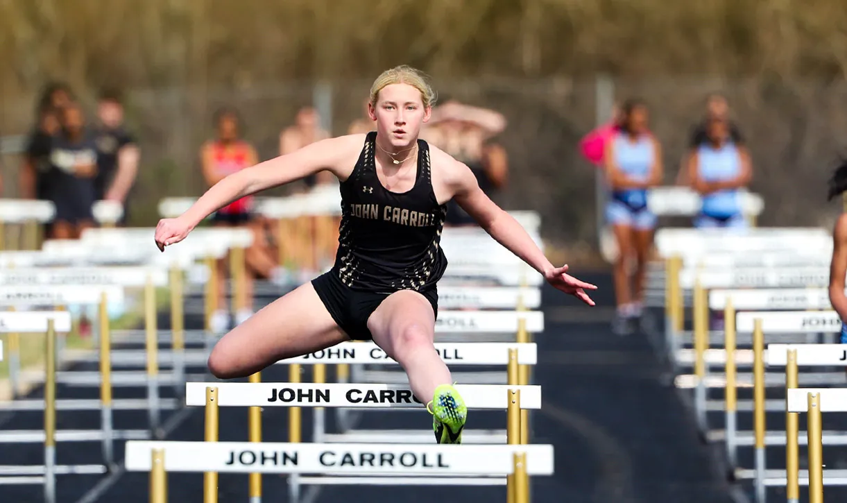 Female athlete in black John Carroll uniform hurdling on an outdoor track during a race.