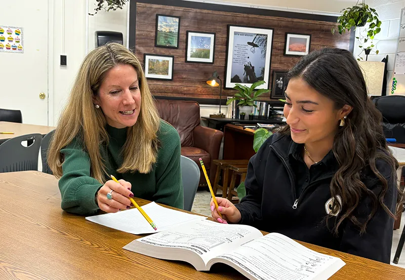 A teacher in a green sweater and a student with dark hair review a workbook together at a classroom table.