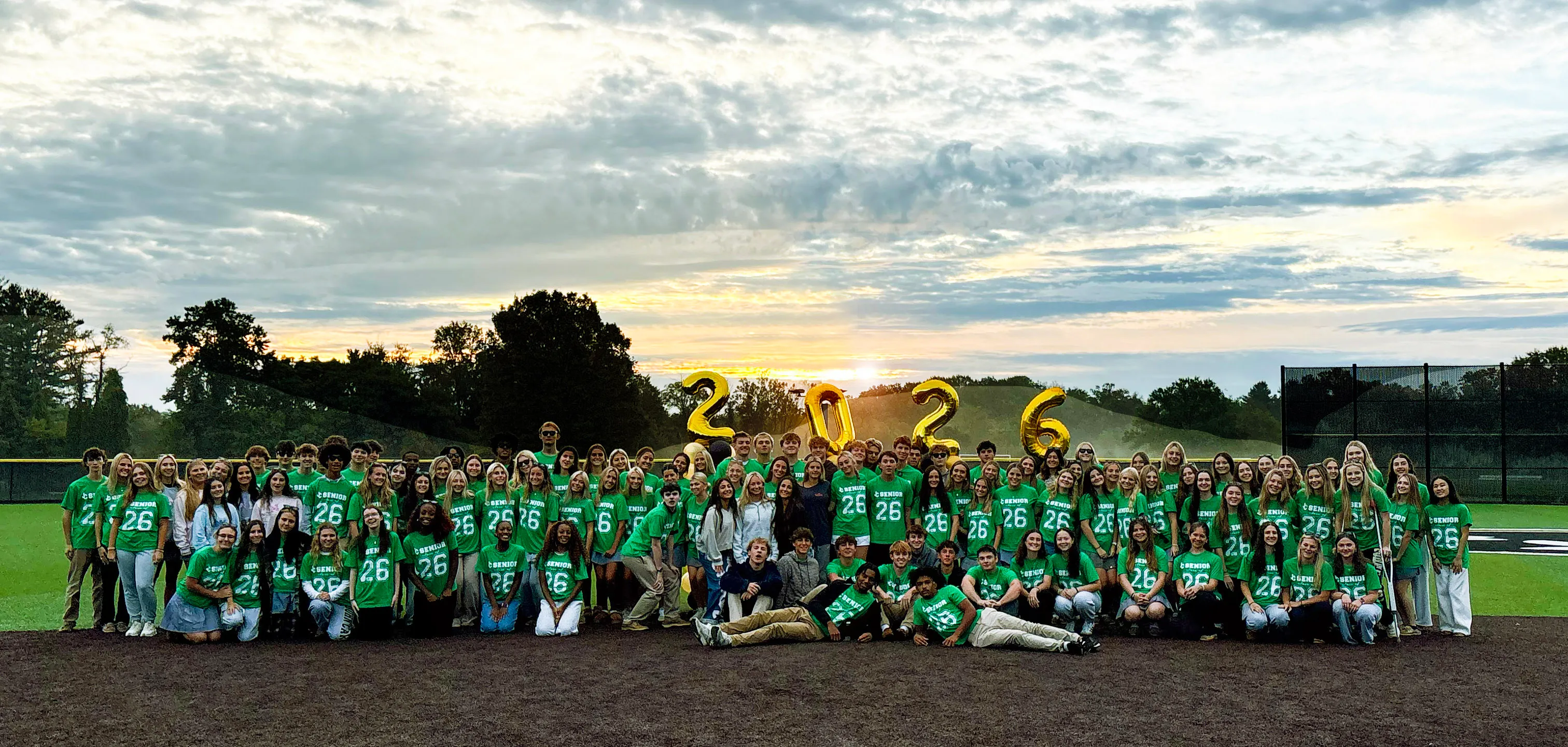 Large group of high school seniors wearing green shirts with 'Senior 26' standing and sitting on a baseball field during a colorful sunrise, holding gold balloons forming 2026.