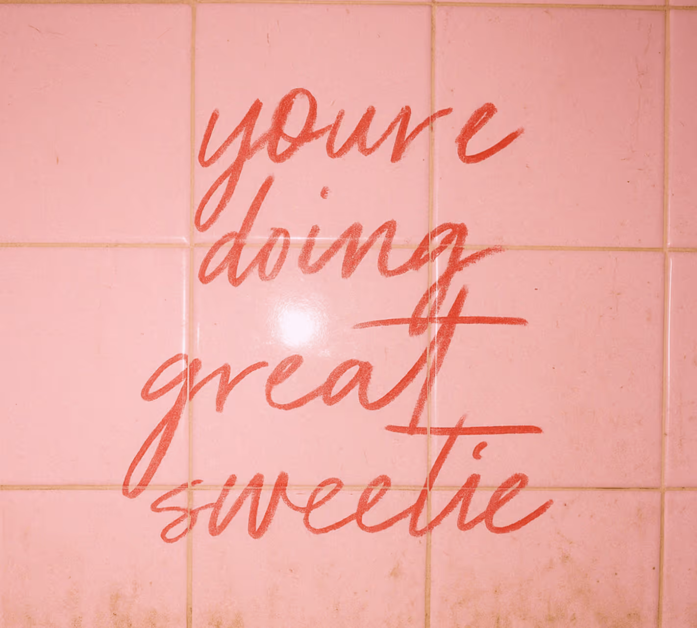 Red handwritten text on pink tiled wall saying, 'you're doing great sweetie'.