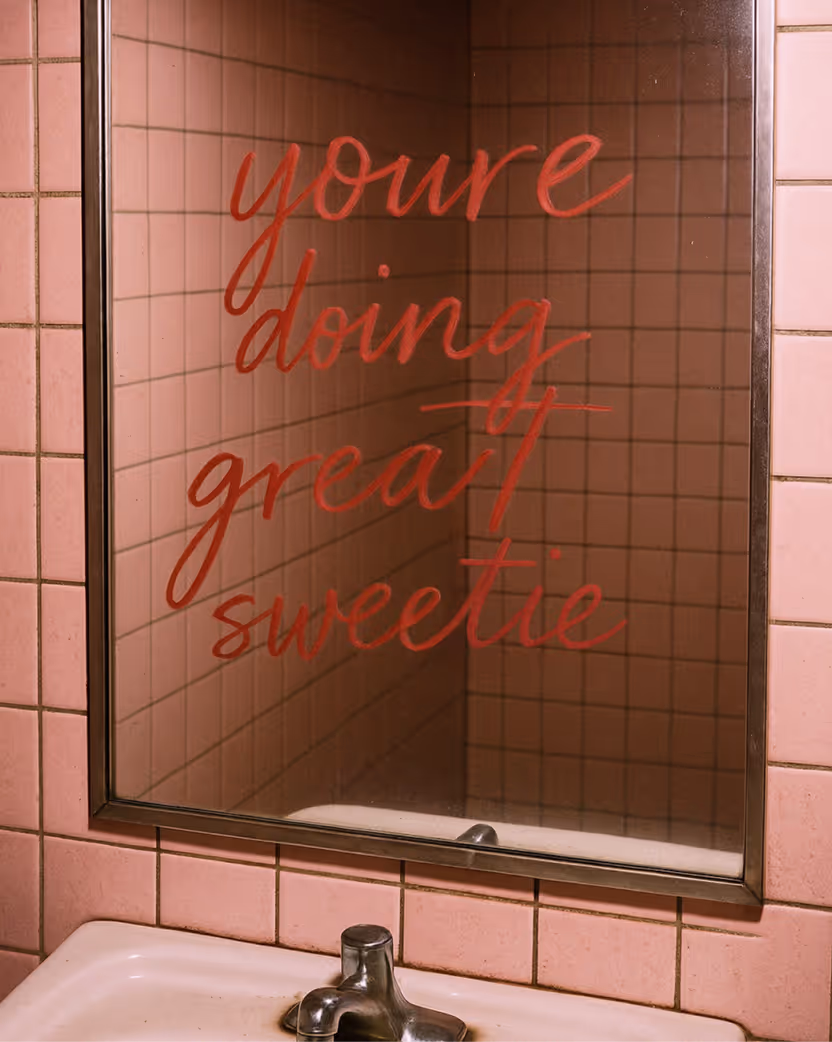 A bathroom mirror with pink tiles reflecting a message written in red lipstick that says "you're doing great sweetie".