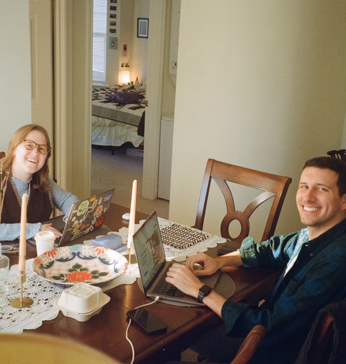 Two founders working on laptops at a dining table in a cozy home environment, decorated with candles and colorful ceramics.