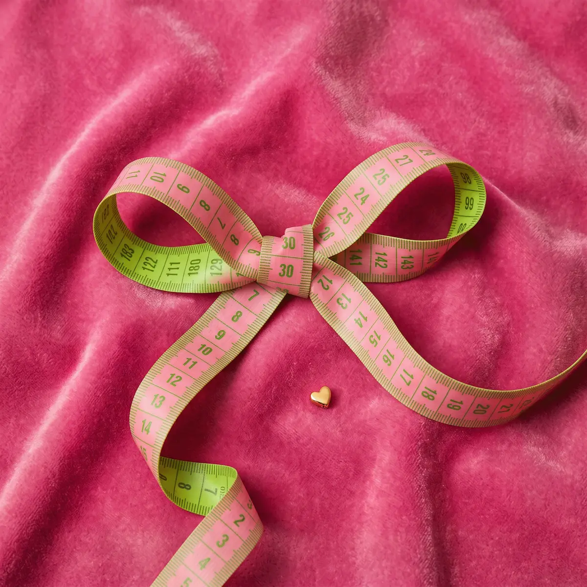Pink measuring tape tied into a bow on pink velvet fabric, with a small gold heart charm
