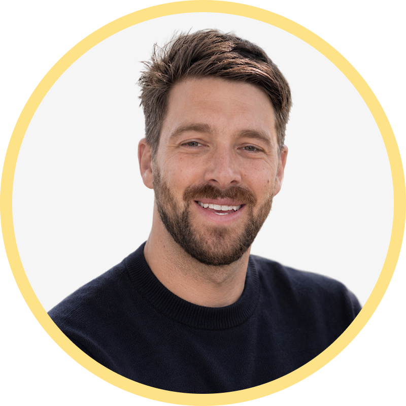 Smiling man with dark hair and beard wearing a black sweater against a plain background.