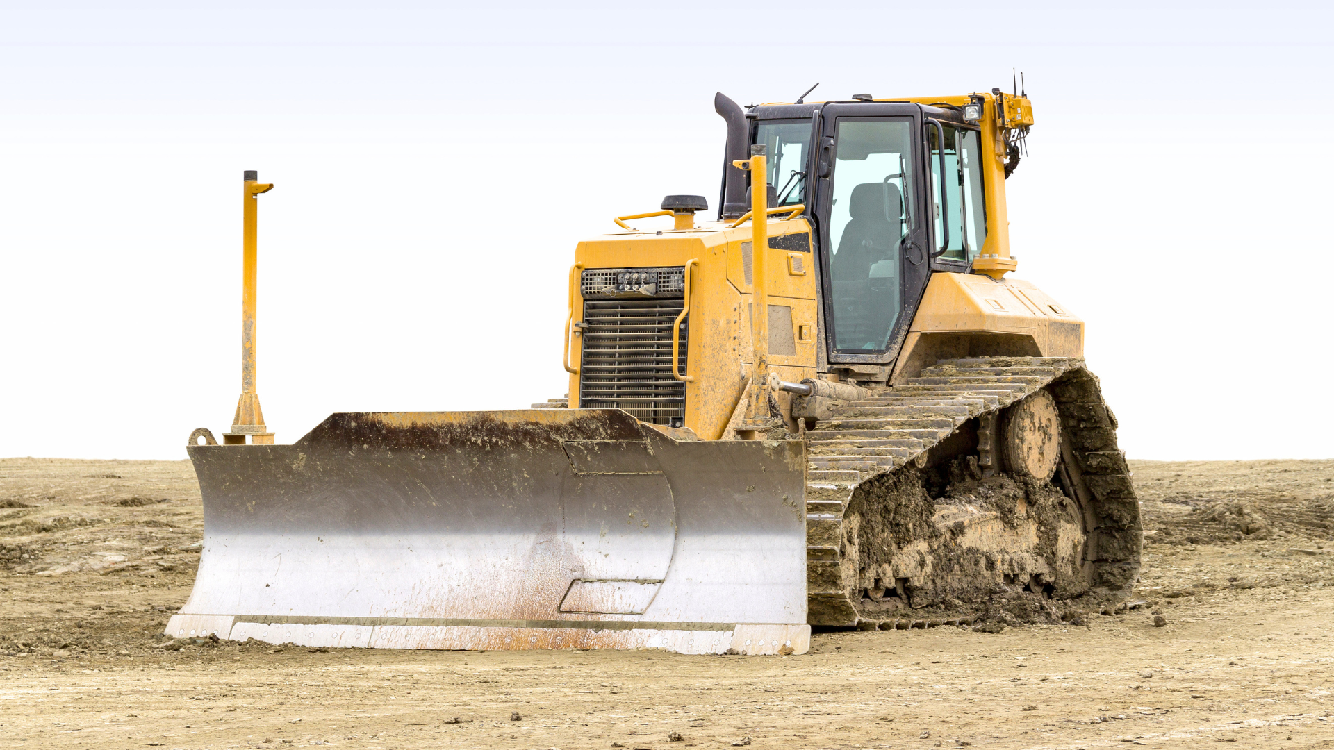 Yellow bulldozer with large blade and tracks parked on dirt ground against white sky.