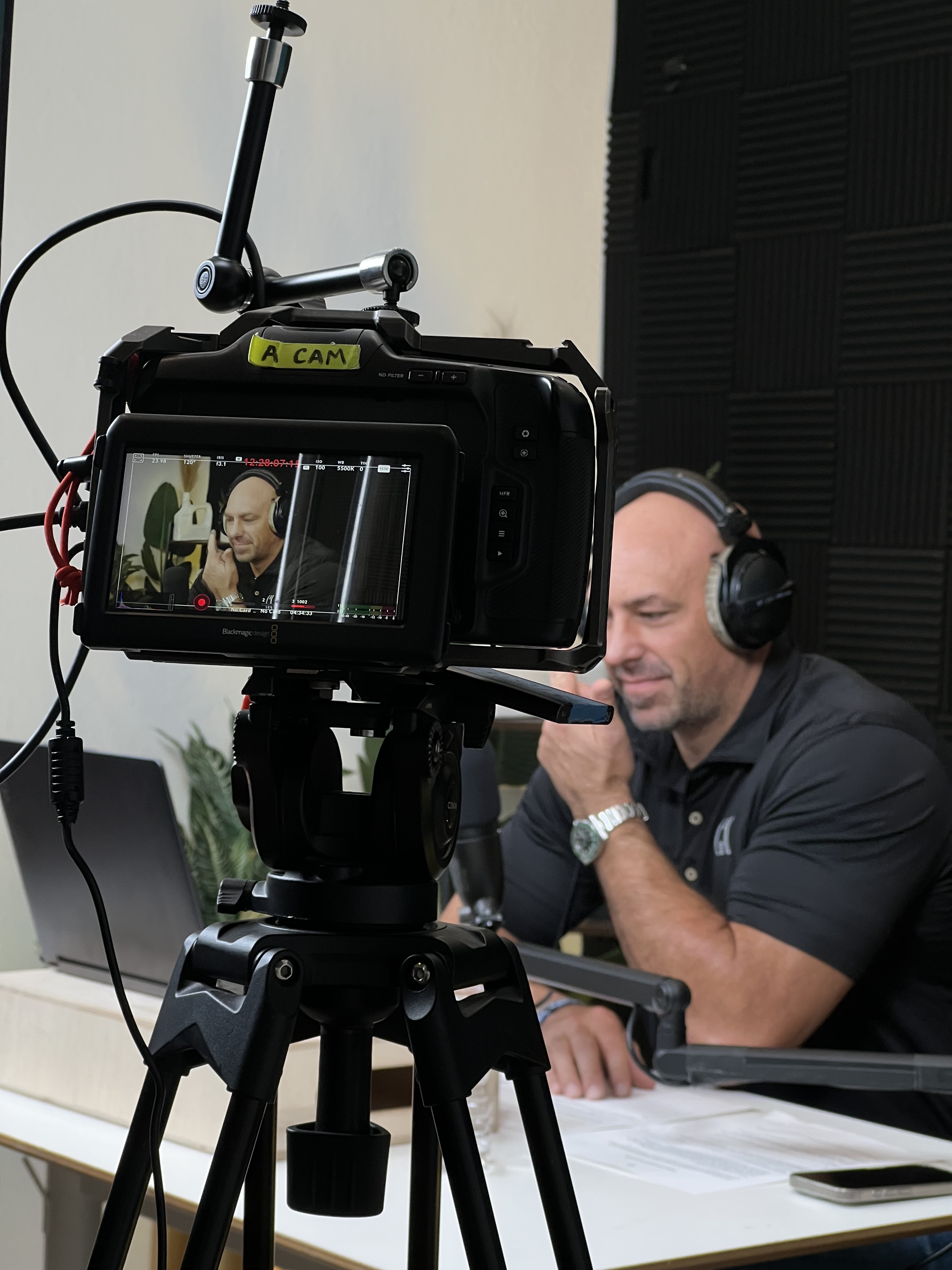 Man wearing headphones speaking into a microphone, recorded by camera on tripod in a studio.