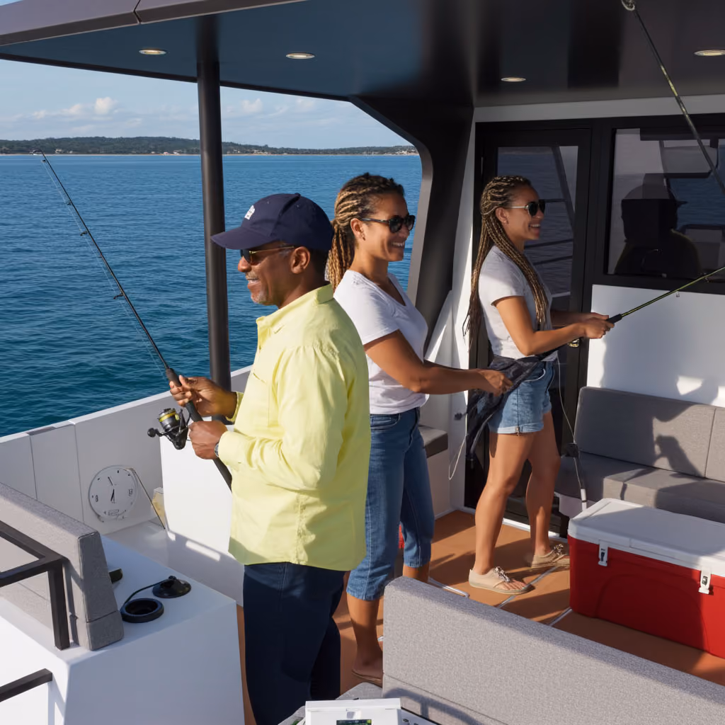 Two women and a man fishing on a boat with calm blue water and a distant shoreline in the background.