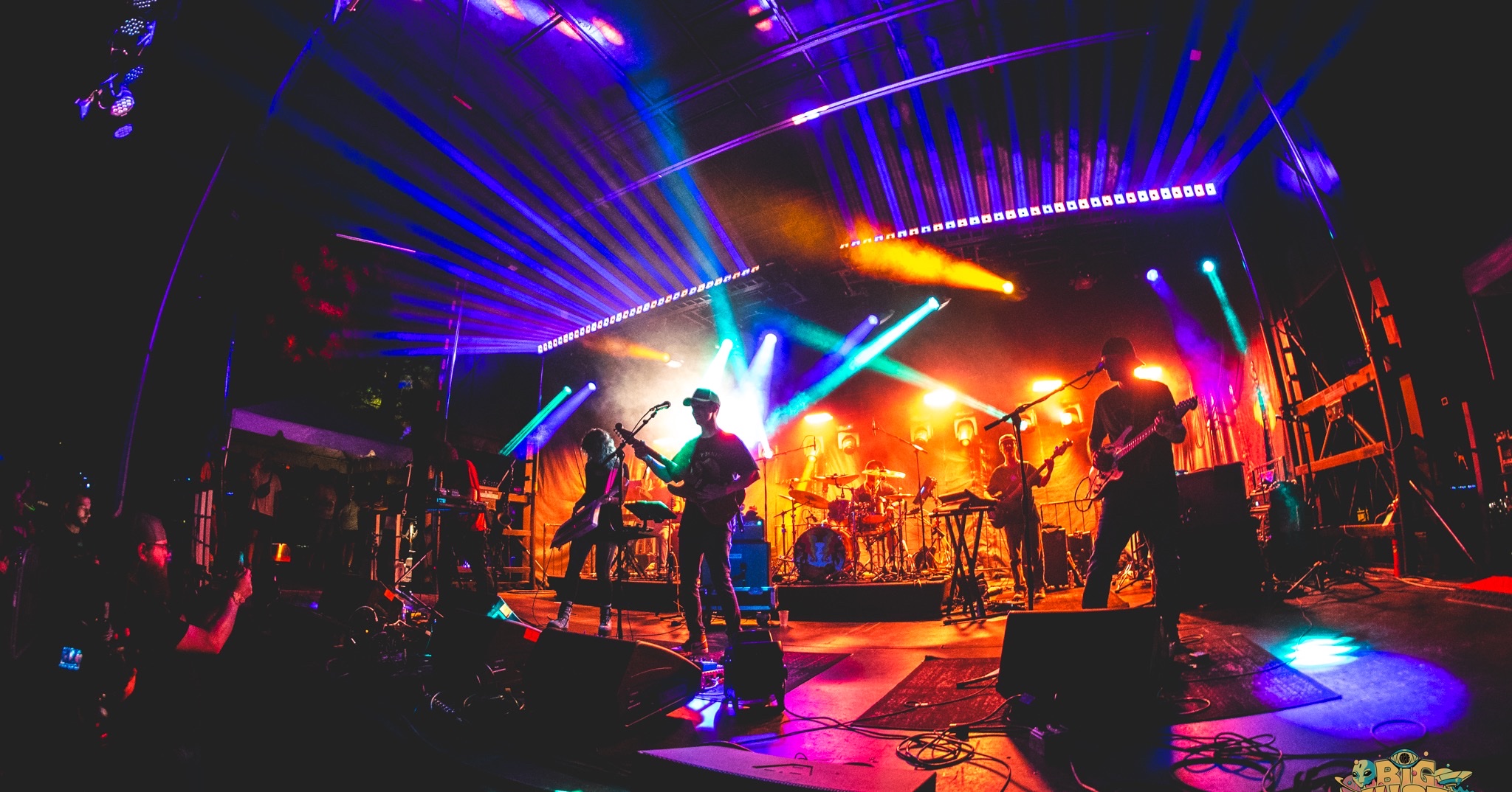 Six piece band on a club stage under blue and violet beams, floor wedges along the deck edge