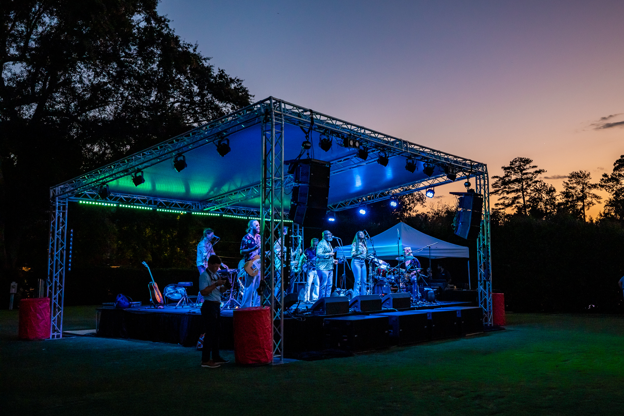 Outdoor band performing on a lit stage at dusk with trees and a colorful sky in the background.