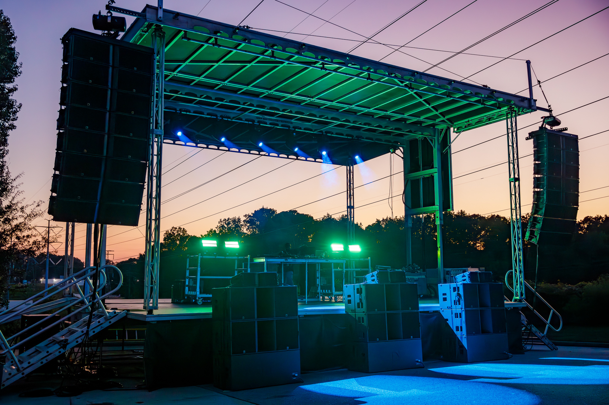 Empty outdoor stage with green and blue lights at sunset, surrounded by trees and large speakers.