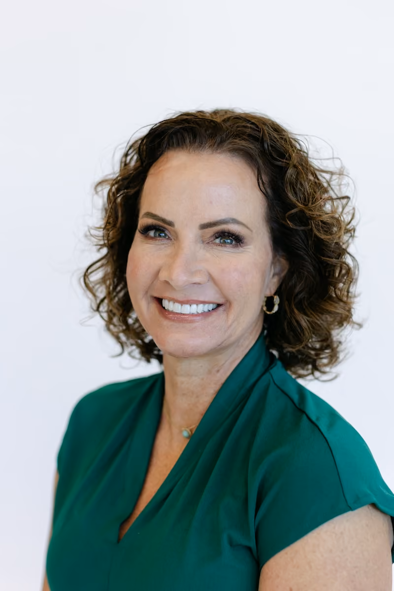 Smiling woman with long blonde hair wearing a white sleeveless top against a plain background.