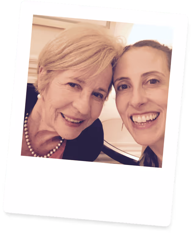 Smiling older woman with short blonde hair and pearl necklace taking a close-up selfie with a younger woman with dark hair.