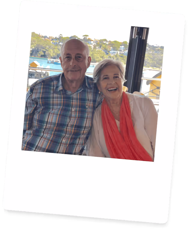 Smiling elderly couple sitting indoors by a large window with a waterfront view in the background.
