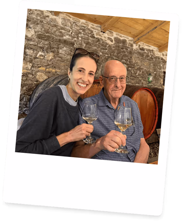 Smiling woman and elderly man sitting together holding glasses of white wine in a rustic stone-walled room.