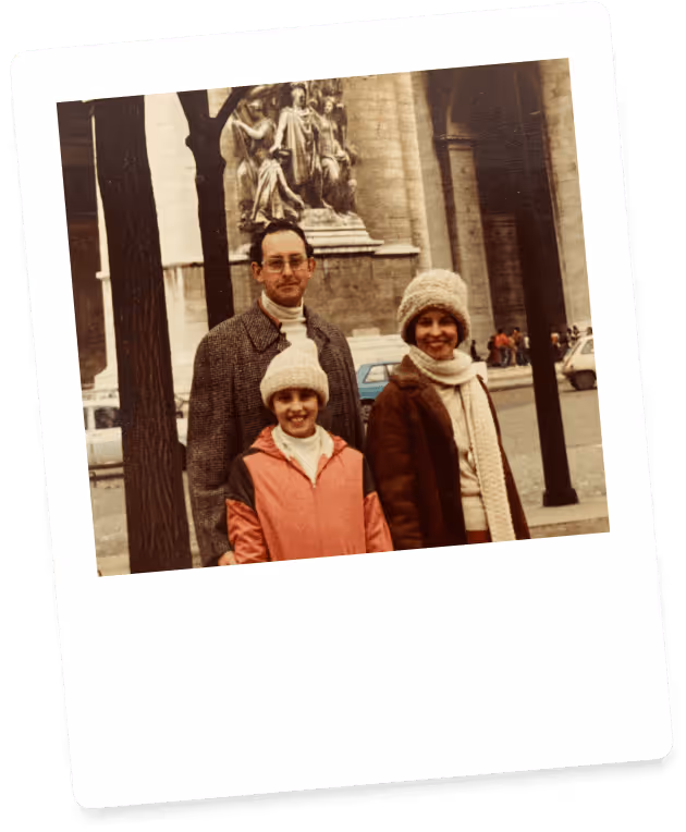 A man, woman, and child wearing winter clothes and knit hats, posing outdoors in front of a historic stone building with sculptures.