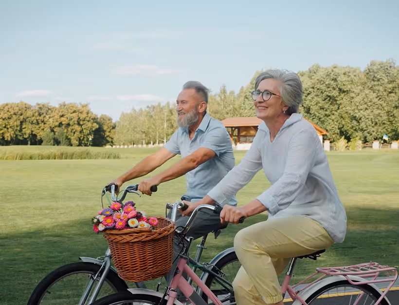 Smiling older couple riding bicycles outdoors on a sunny day with a basket of colorful flowers on the woman's bike.