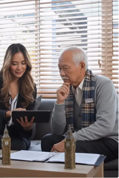 A young woman showing something on a tablet to an elderly man sitting beside her with window blinds in the background.