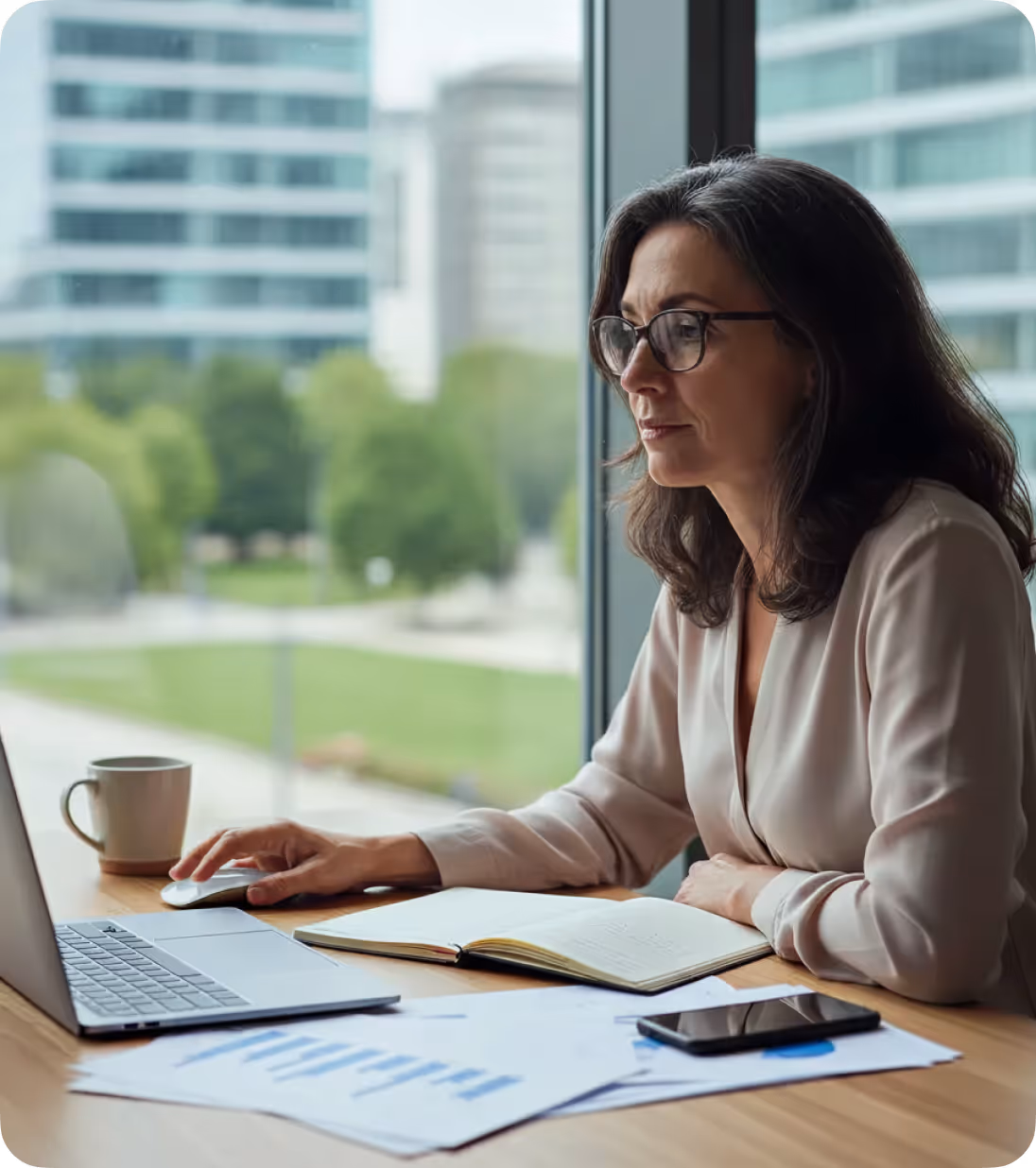 Woman with glasses sitting at a desk by a window, working on a laptop with documents and a notebook nearby.