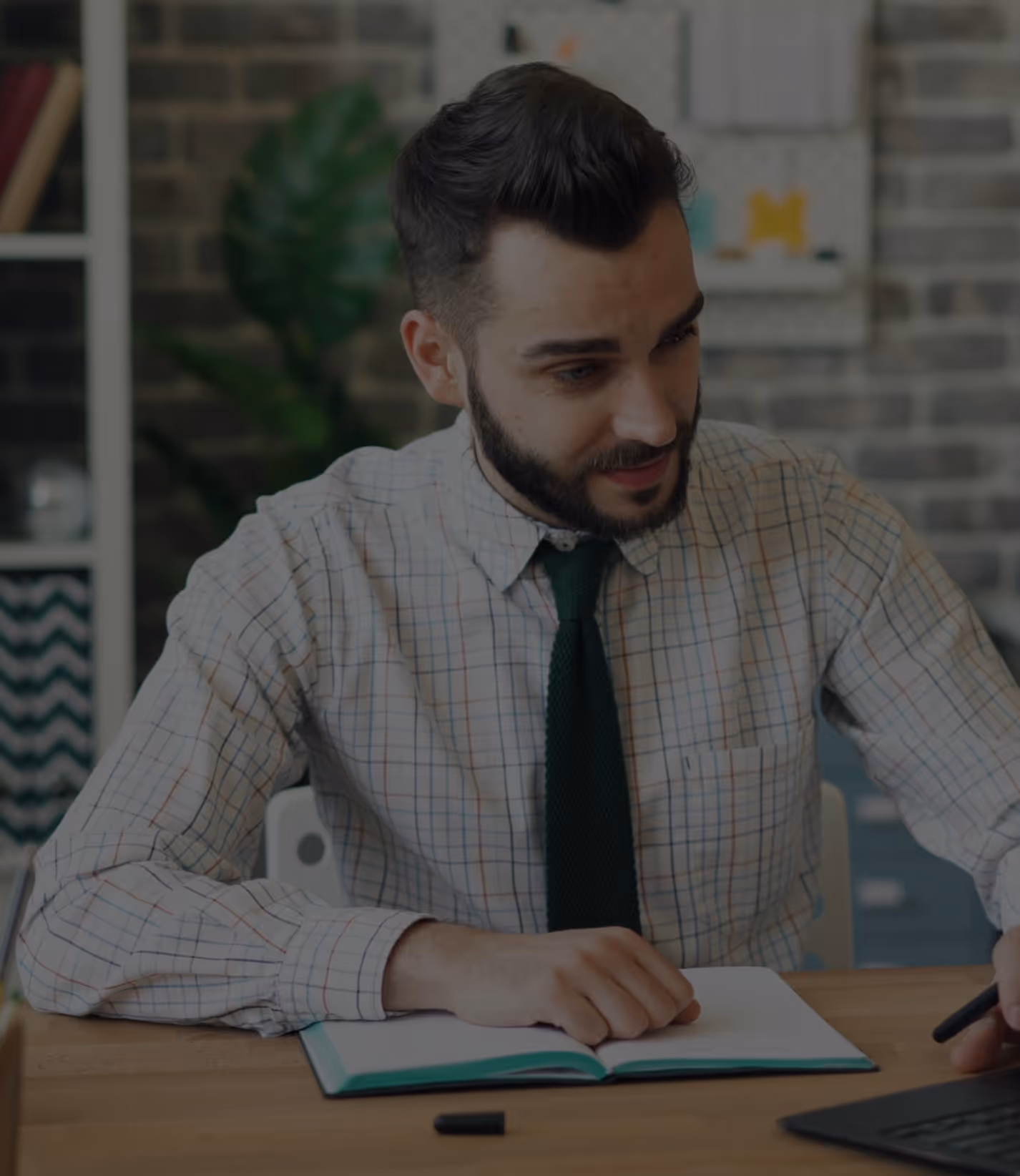Man in a checked shirt and green tie sitting at a desk with an open notebook and a laptop.