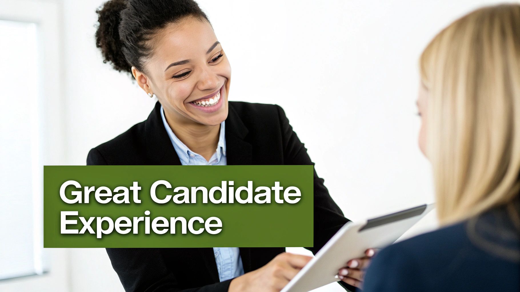 A smiling woman in a suit engages with another professional holding a tablet, suggesting a positive candidate experience.