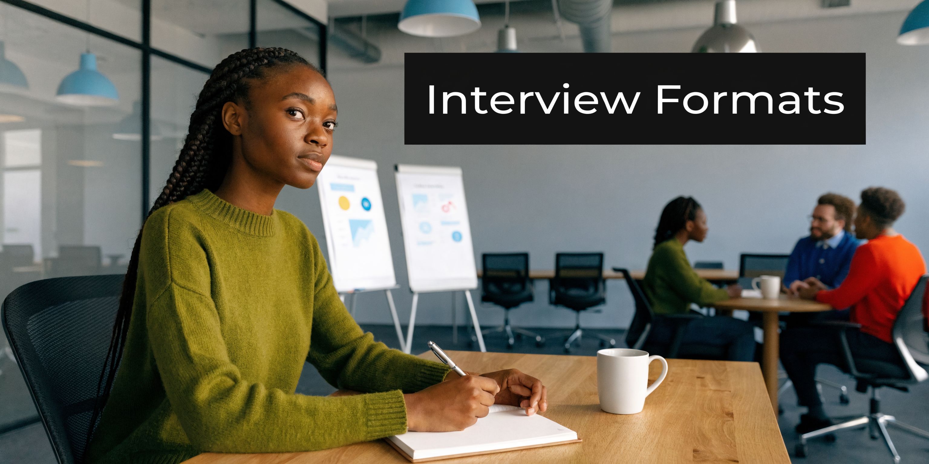 A young woman sits at a desk in a modern office writing in a notebook while others meet.