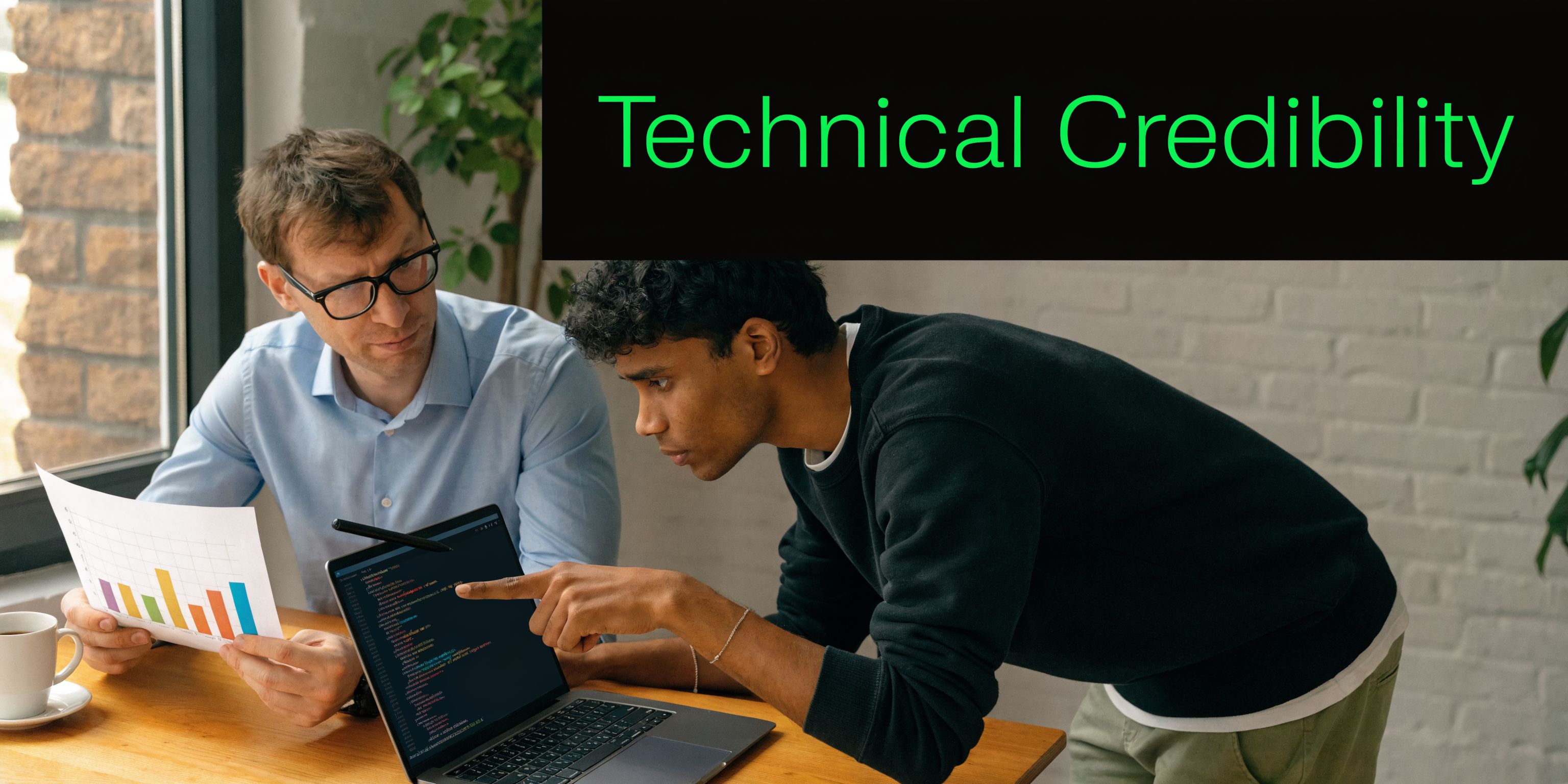 A manager and a developer collaborating over code on a laptop and reviewing a bar chart document.