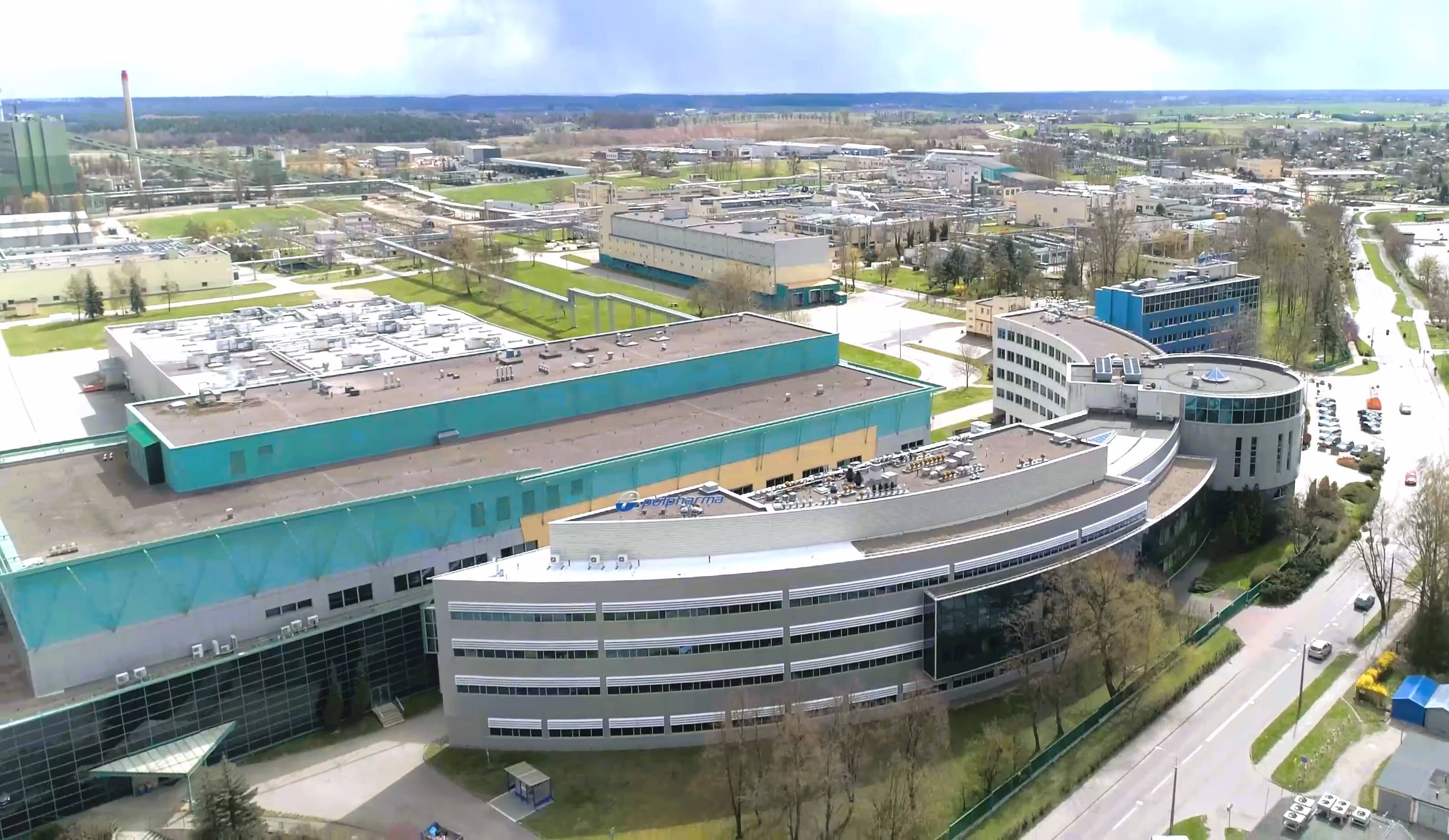 Aerial view of a large industrial complex with multiple modern buildings, surrounded by trees and roads on a clear day.