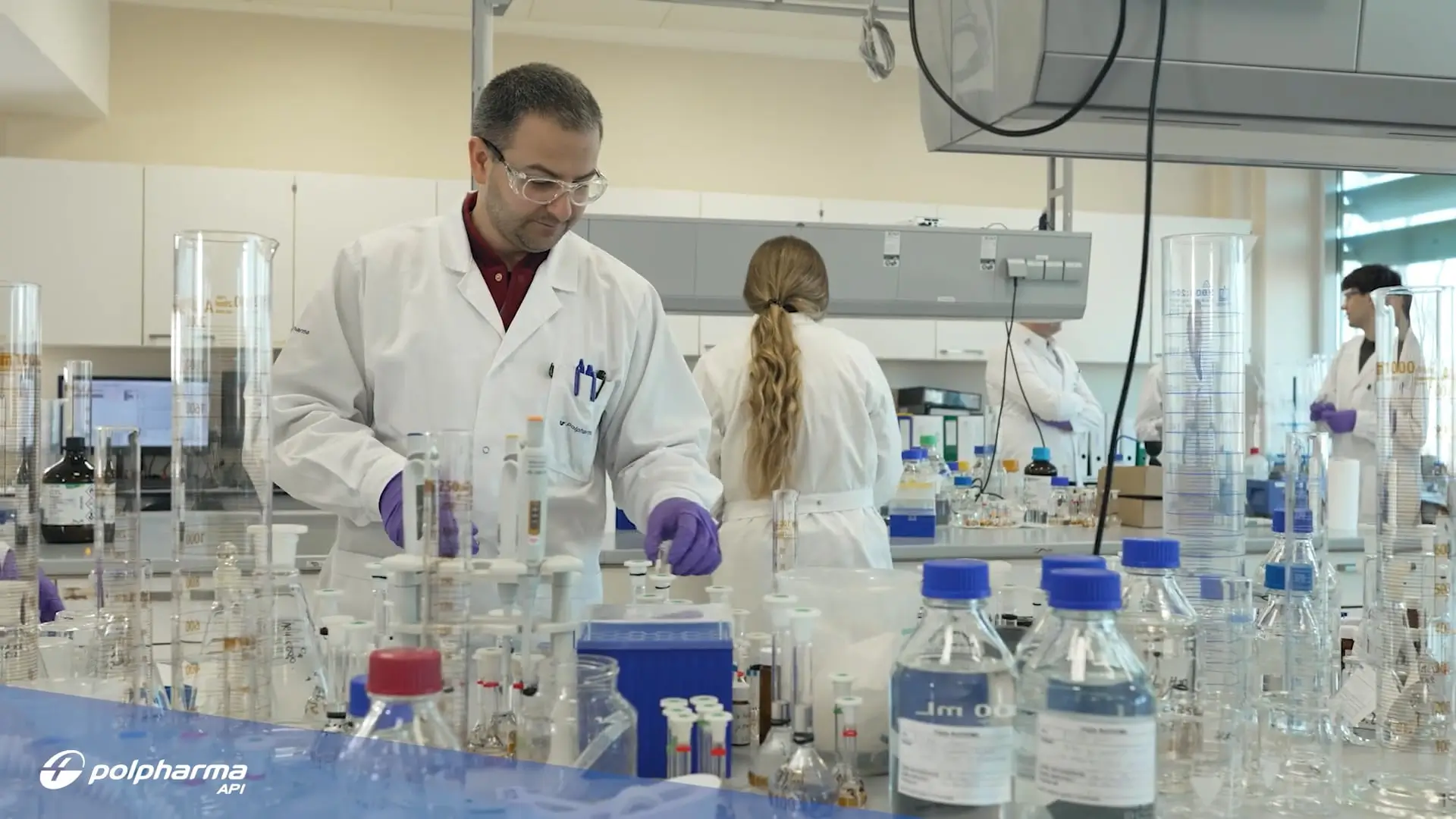 Scientists in white lab coats and purple gloves working with glassware and chemical bottles in a modern laboratory setting with Polpharma API logo in the corner.