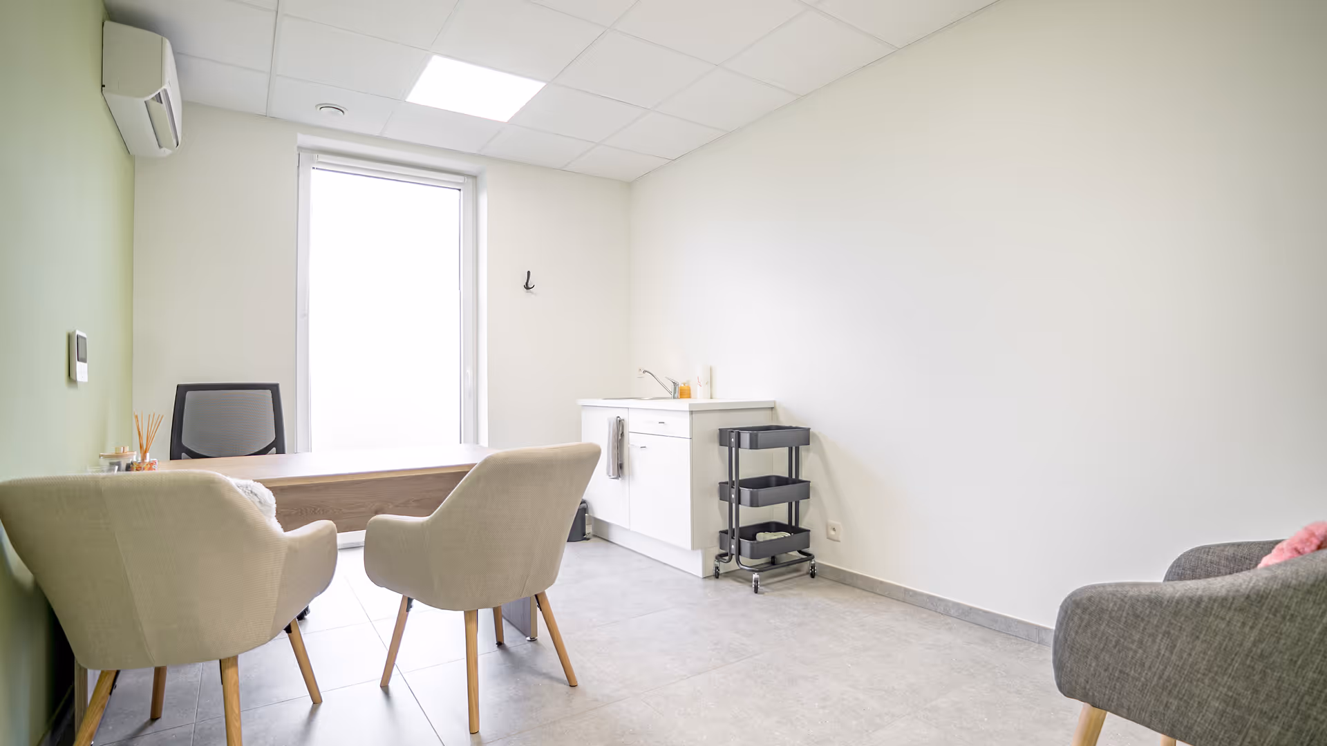 Bureau lumineux avec deux fauteuils beige, un bureau en bois, une chaise de bureau noire, un meuble blanc avec un évier et un chariot à roulettes noir.