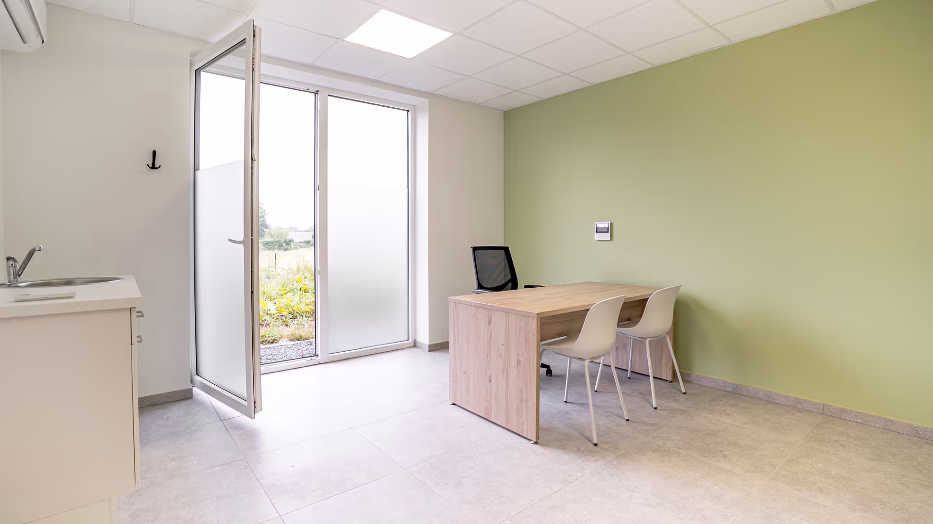 Bureau minimaliste avec une table en bois clair, deux chaises blanches et une chaise noire, à côté d'une porte-fenêtre ouverte donnant sur un jardin.