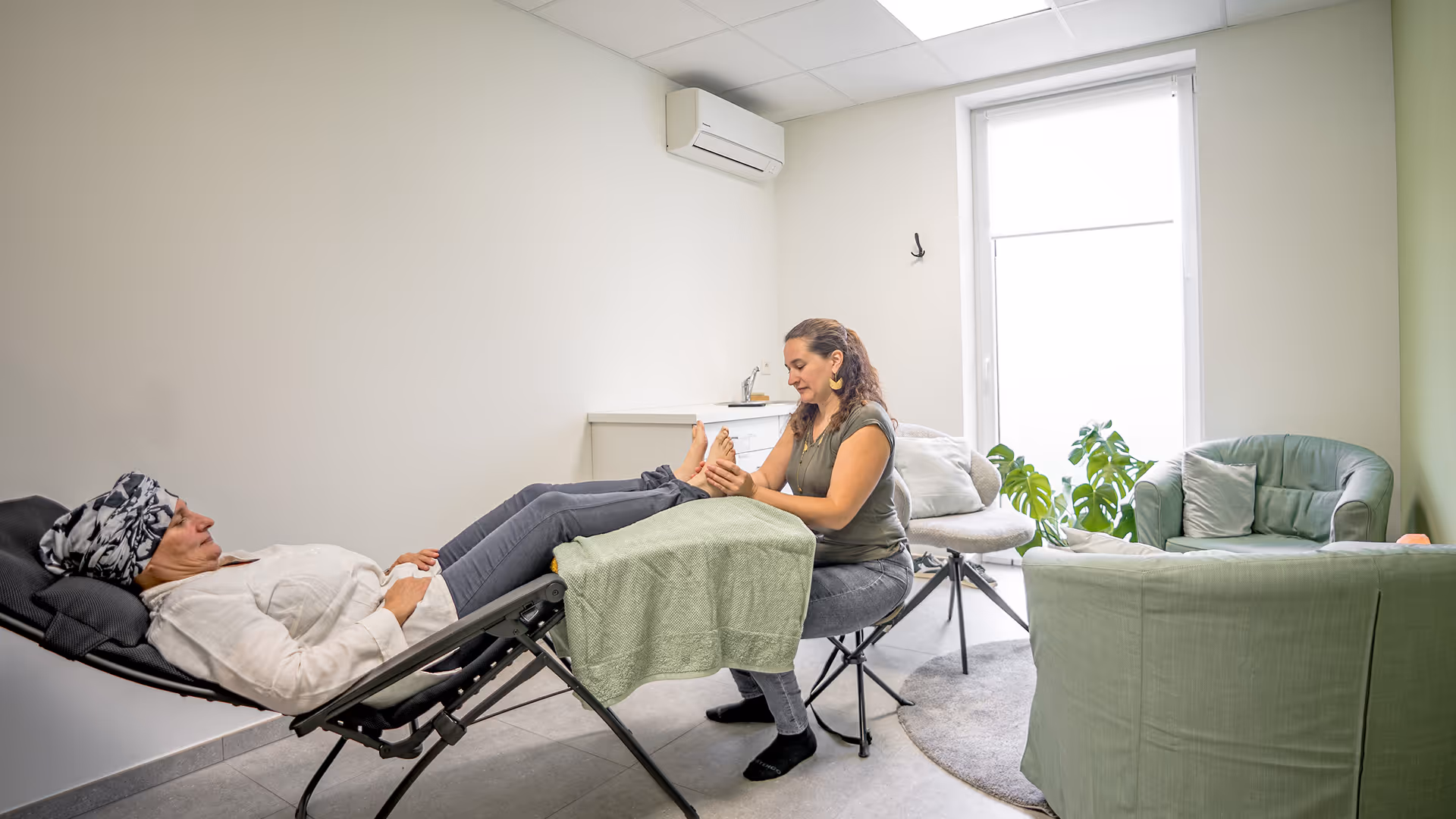 Une thérapeute masse les pieds d'une patiente allongée sur une chaise longue noire dans une pièce lumineuse avec des fauteuils verts et une plante décorative.