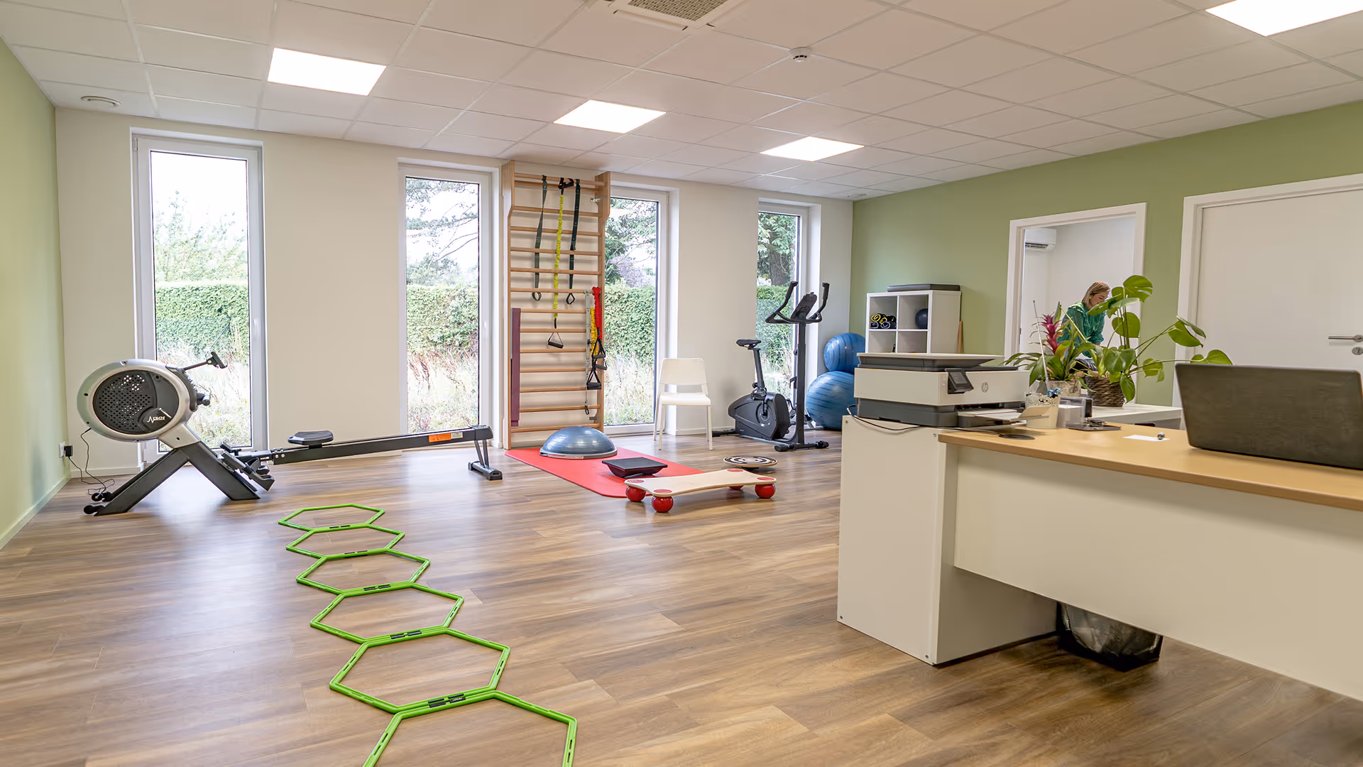 Salle de physiothérapie lumineuse avec équipements d'exercice, tapis rouge, vélo d'appartement, et un bureau avec une plante verte.