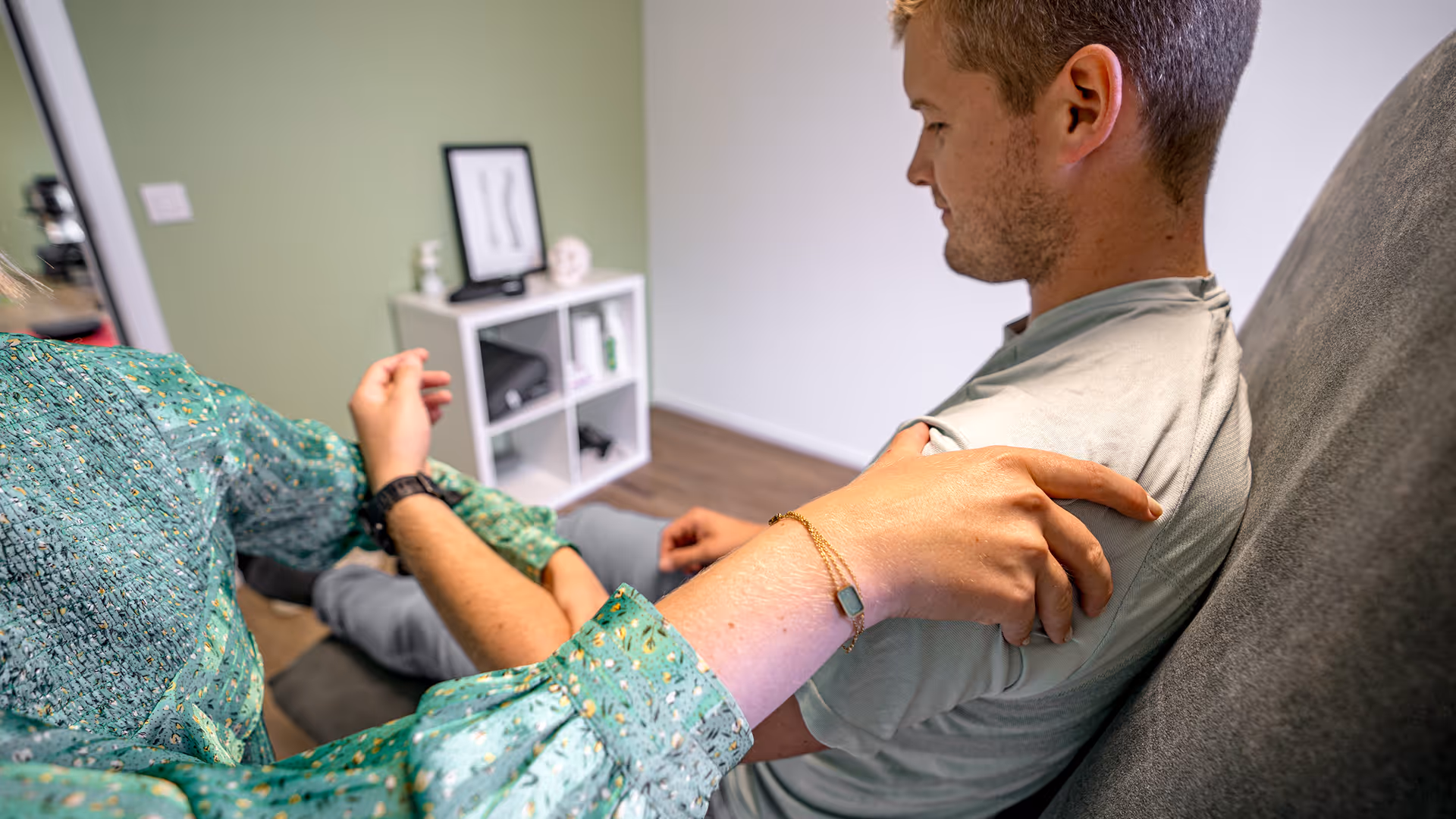 Une femme en blouse verte touche doucement l'épaule d'un homme assis à côté d'elle.