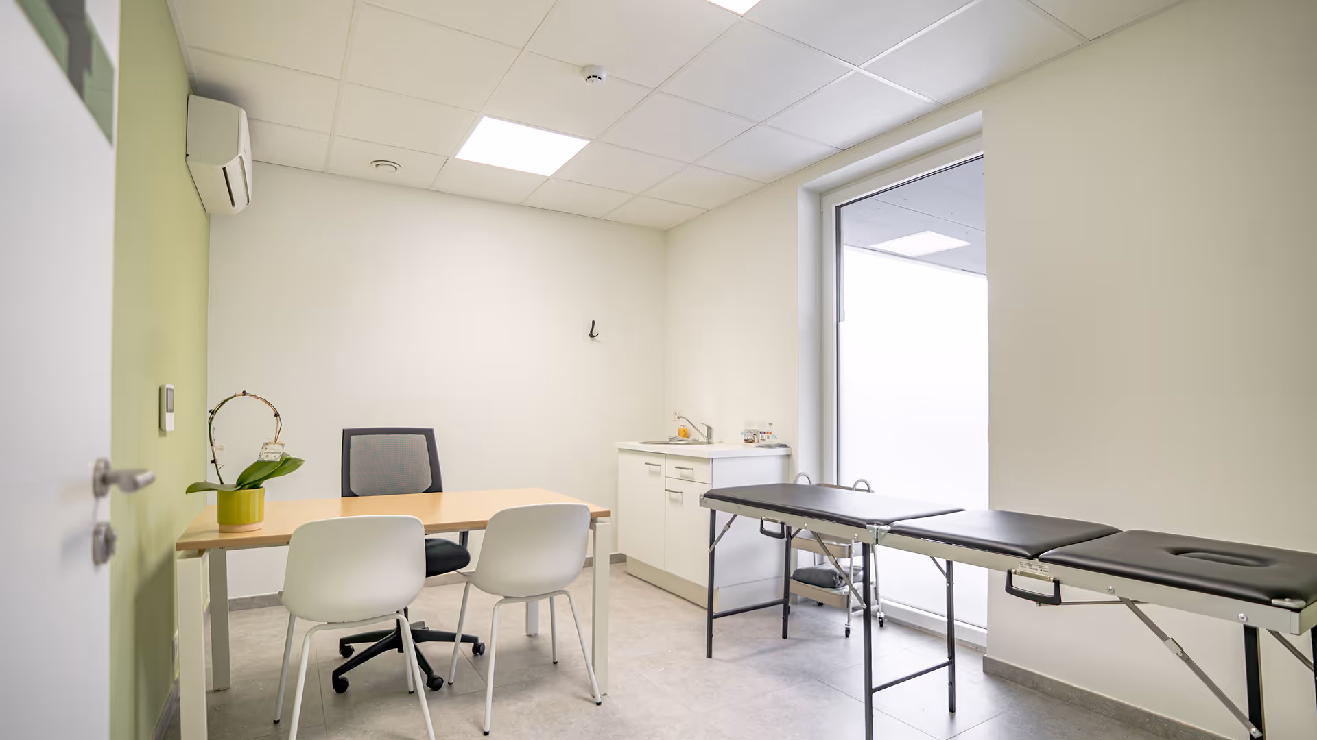 Cabinet médical épuré avec chaise de bureau, deux chaises blanches, table et table d'examen noire près d'une grande fenêtre.