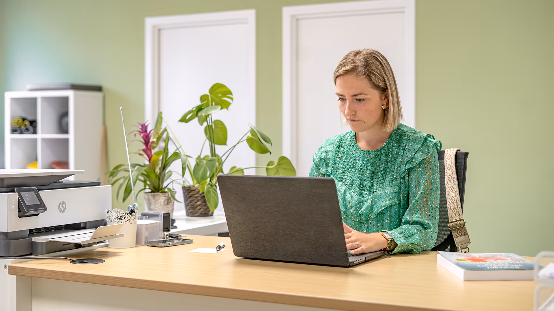 Femme blonde portant un chemisier vert travaillant sur ordinateur portable dans un bureau clair avec des plantes et une imprimante sur le bureau.