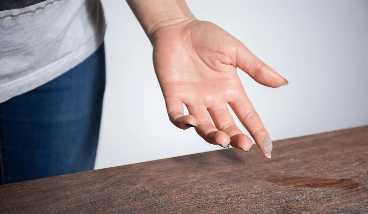 Close-up of a woman's finger showing a thick layer of dust accumulated on it after swiping it across a dusty table, highlighting the need for post-construction cleaning in a room with visible construction debris and tools in the background.