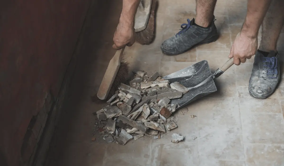 A man sweeping construction debris with a large broom in a room filled with remnants of building materials such as wood scraps and plaster. The image captures the man in action, emphasizing the thorough clean-up process required after construction work.