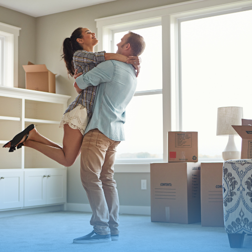 A joyful couple smiling as they unpack boxes and arrange decor in their new home, with sunlight streaming through the windows, signifying a fresh start in their bright and airy living space.