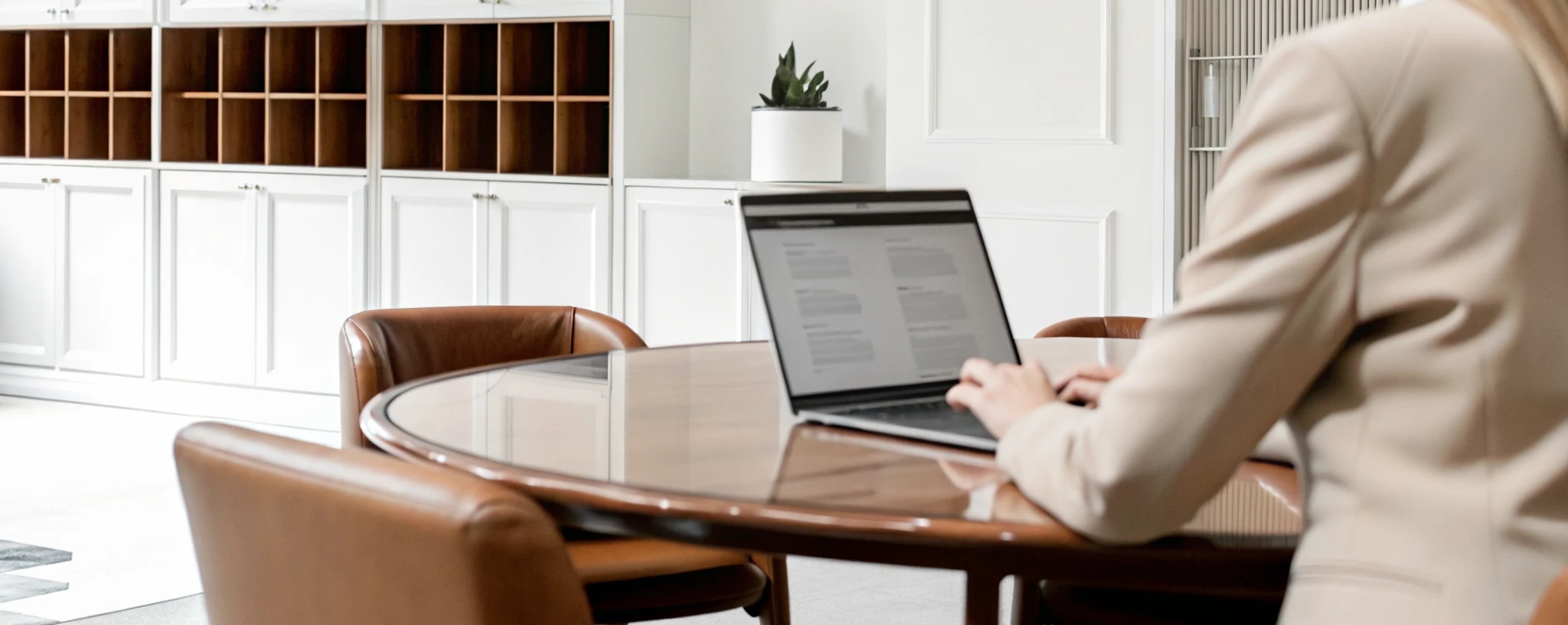 A person seated at a round wooden table working on a laptop in a bright, modern office with white cabinets, open shelving, and neutral-toned furniture.
