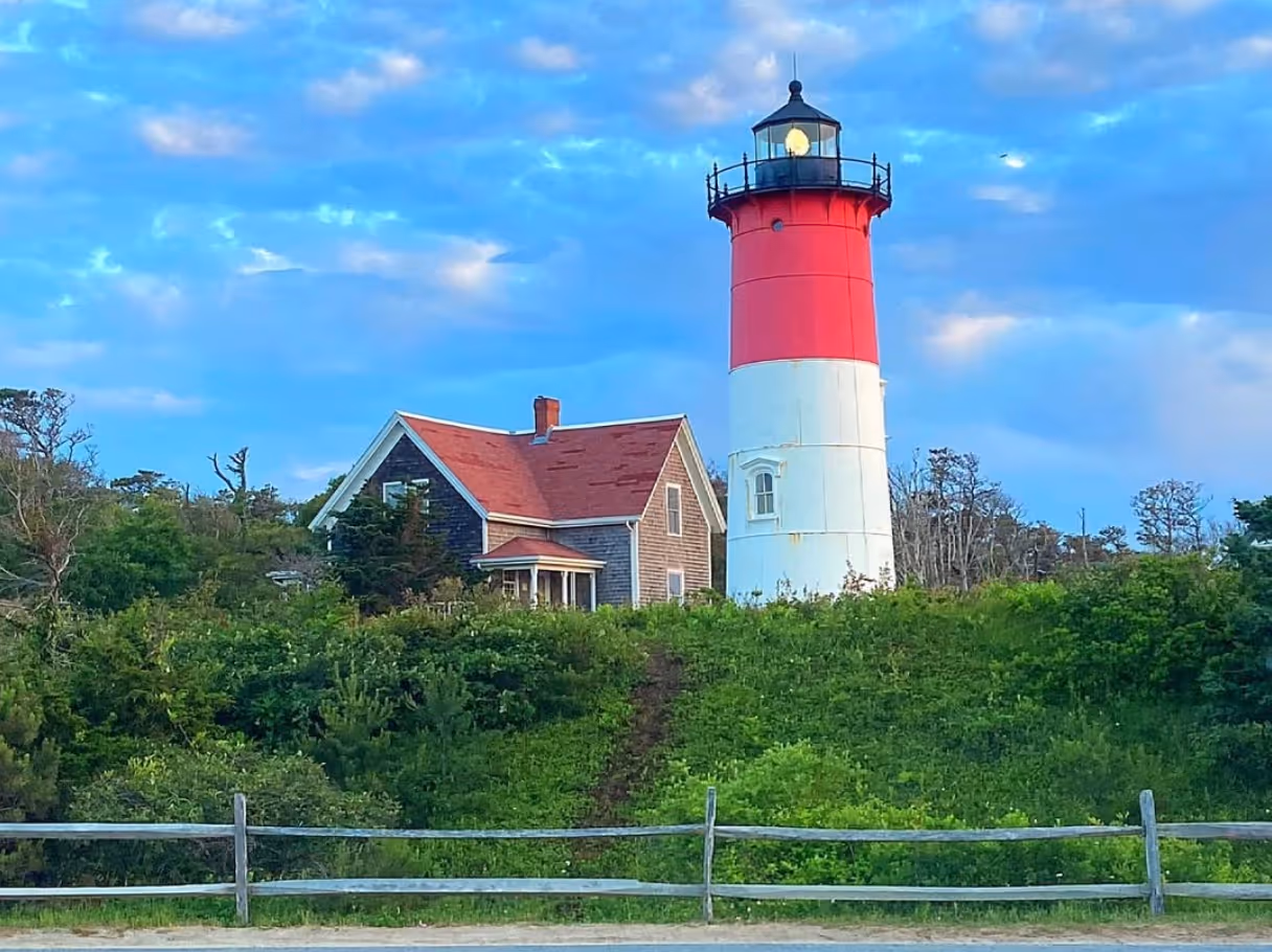 A light house in Eastham MA during the day rising above the tree-line. A blue summer sky sits atop the photo.