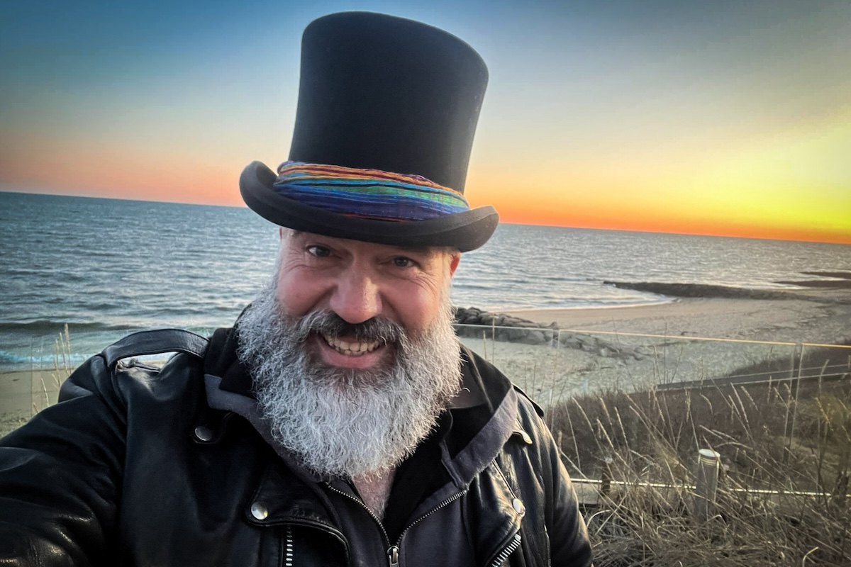 Derek Dibbern on a Cape Cod beach in front of sunset with his top hat and pronounced beard.