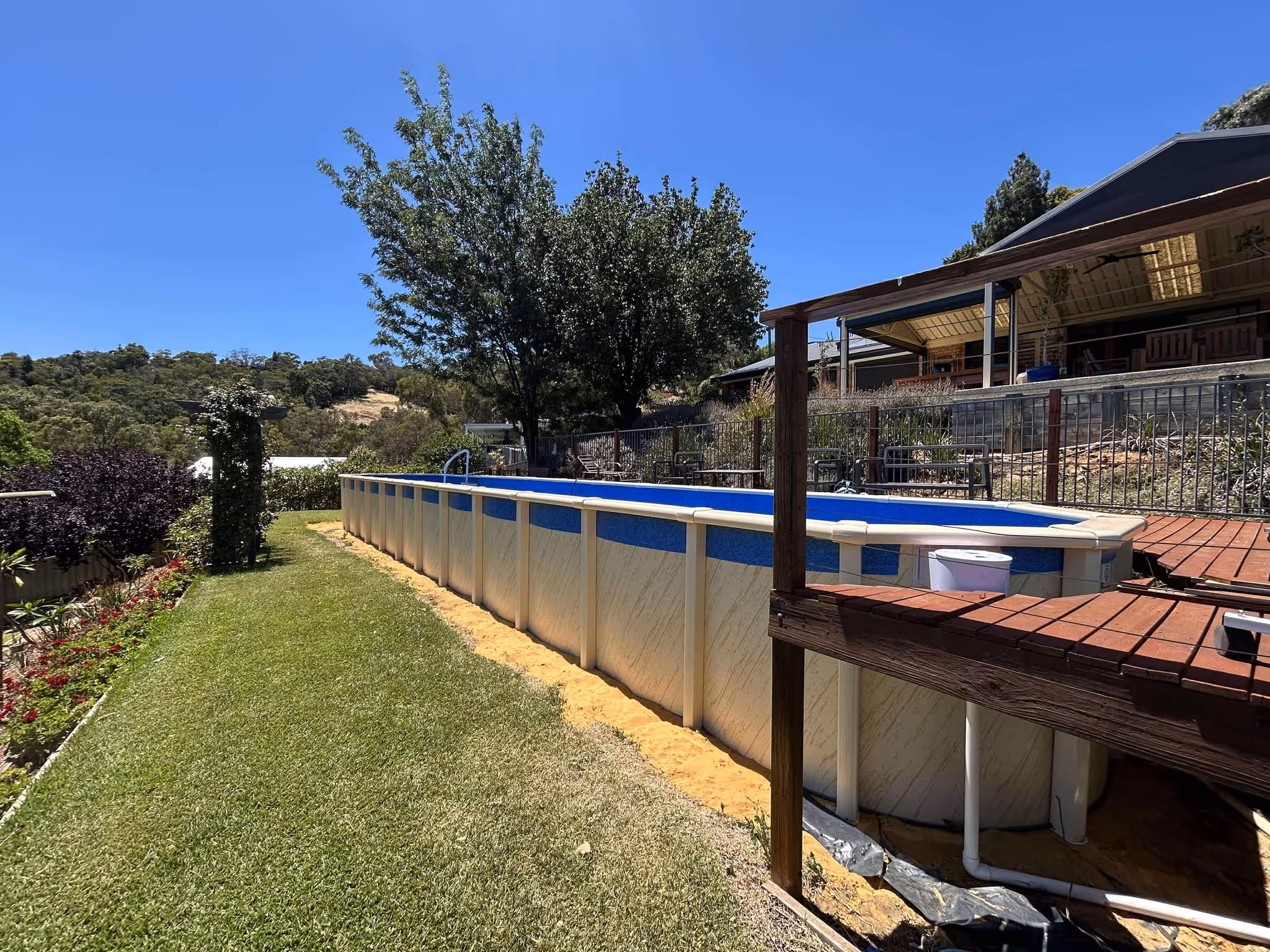 Above-ground oval pool installed on a sloped lawn, next to a wooden deck and garden with hill views.