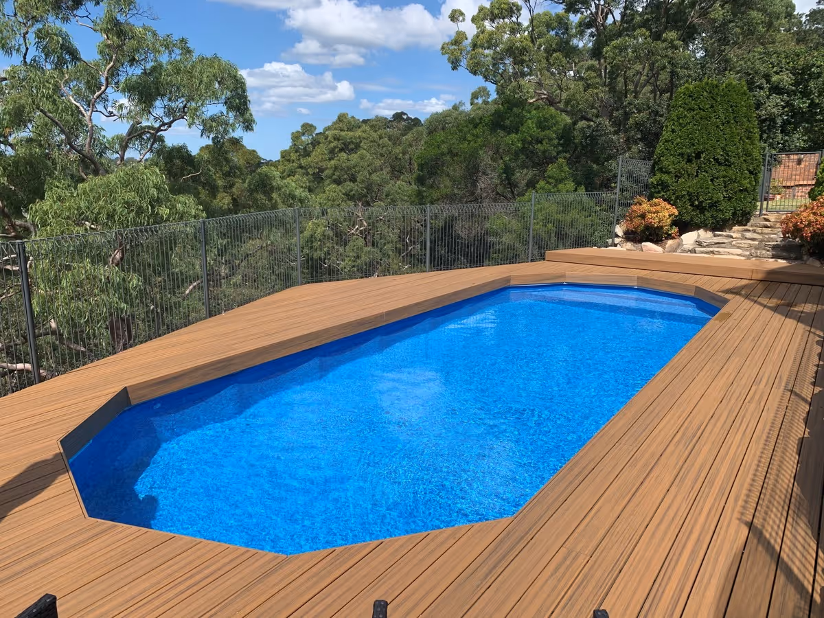 Rectangular blue swimming pool surrounded by brown wooden deck and green trees under a partly cloudy sky.