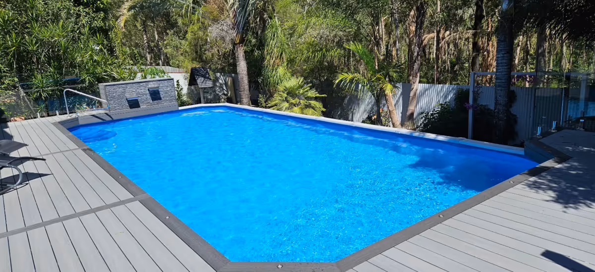 Outdoor rectangular swimming pool with bright blue water surrounded by gray deck and tropical plants.