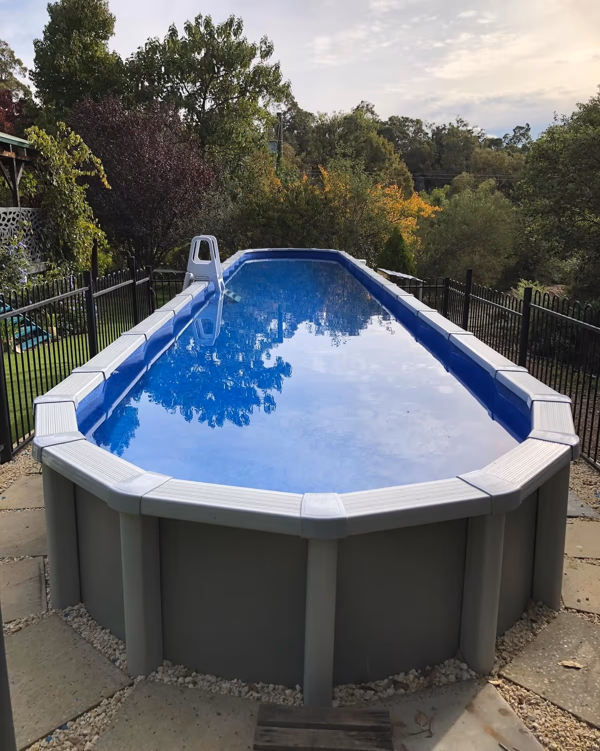 Above ground rectangular swimming pool with clear water reflecting trees and sky, surrounded by garden and fencing.