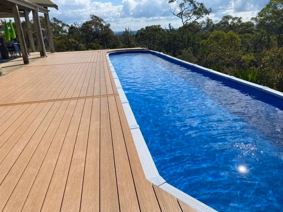 Long outdoor swimming pool with clear blue water beside a wooden deck with trees in the background.