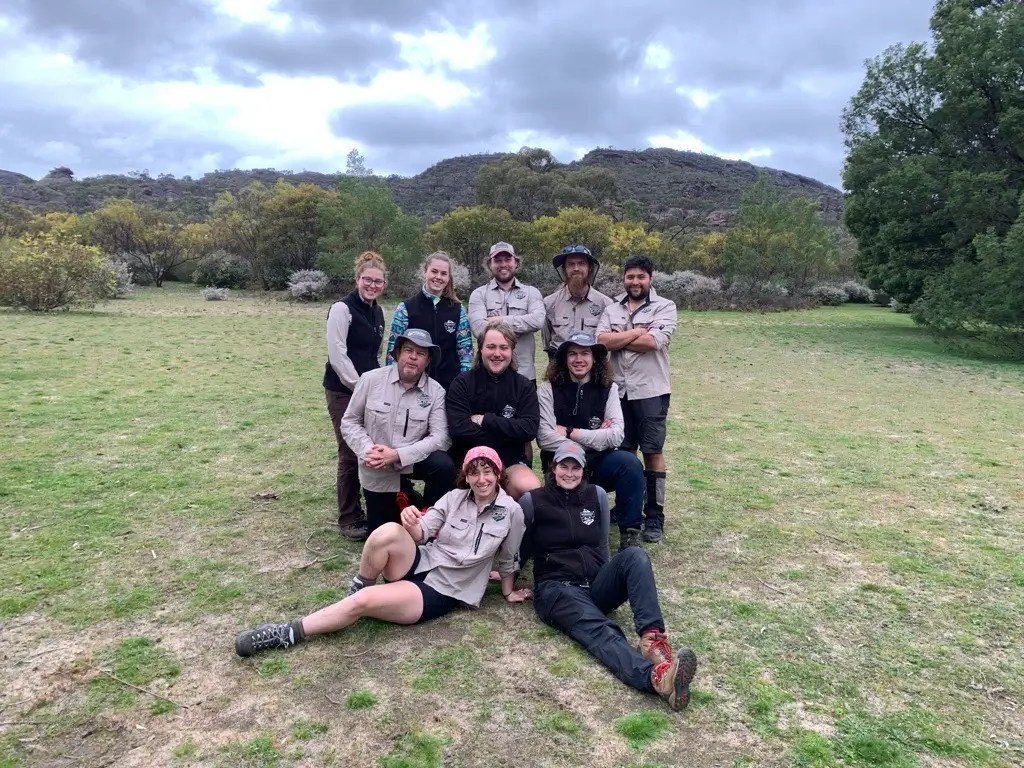 A group of ten outdoor staff posing in coordinated uniforms on grassy terrain with hills and cloudy skies in the background.