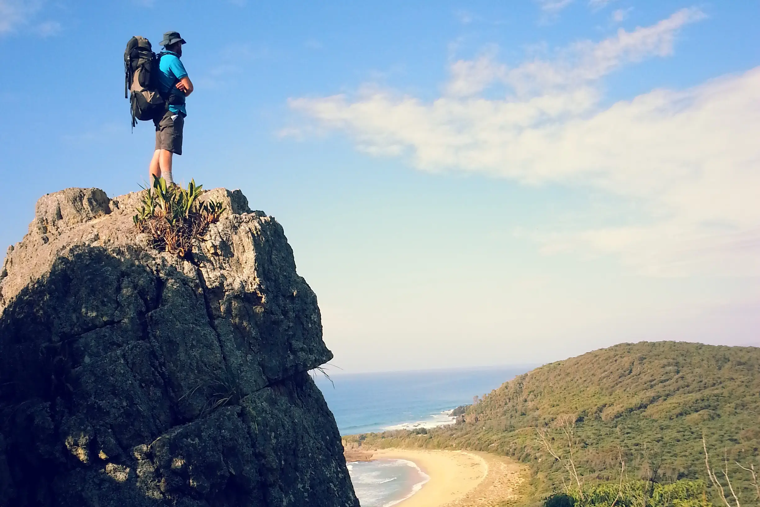 A hiker with backpack standing atop a rocky summit overlooking a coastal beach and forested hillside beneath a blue sky.
