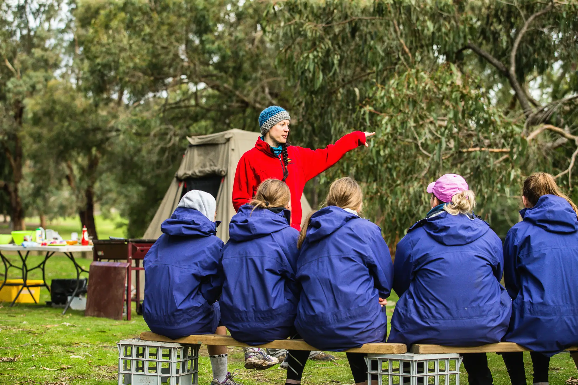 An instructor in a red jacket gesturing whilst teaching a group of students in matching blue hoodies seated outdoors at a campsite.
